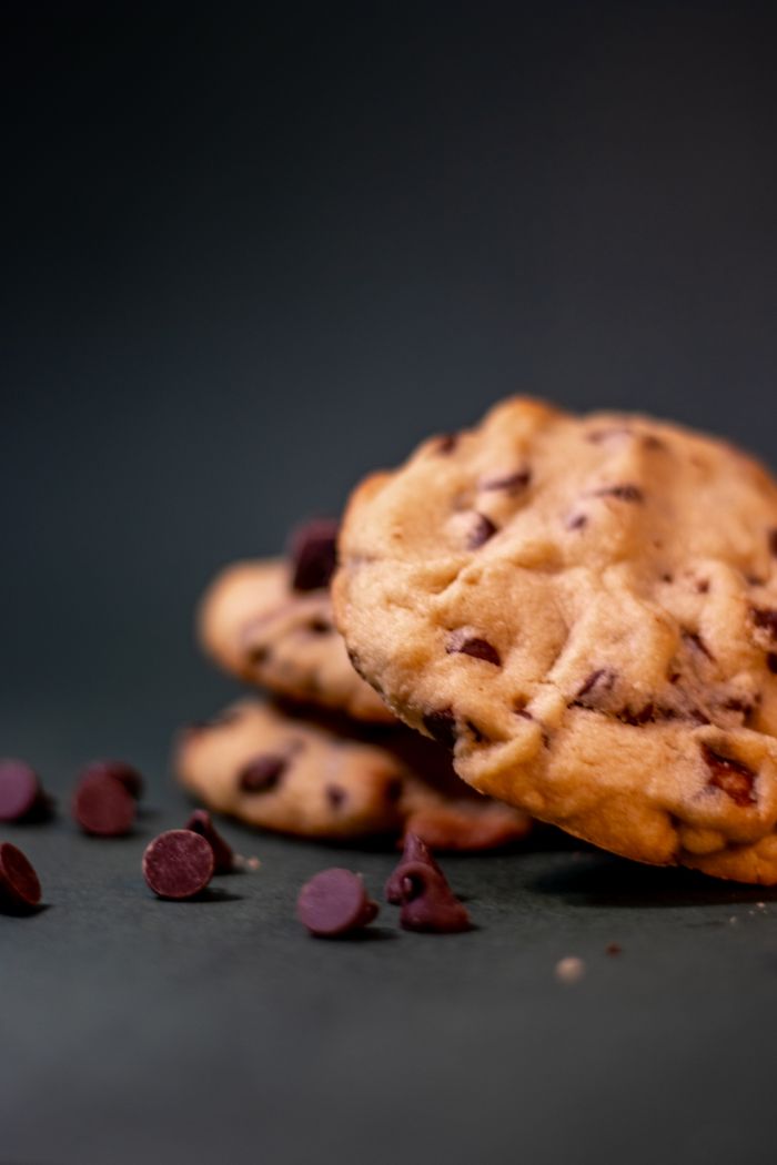 about-01 Close-up of stacked chocolate chip cookies with scattered chips on a dark background.